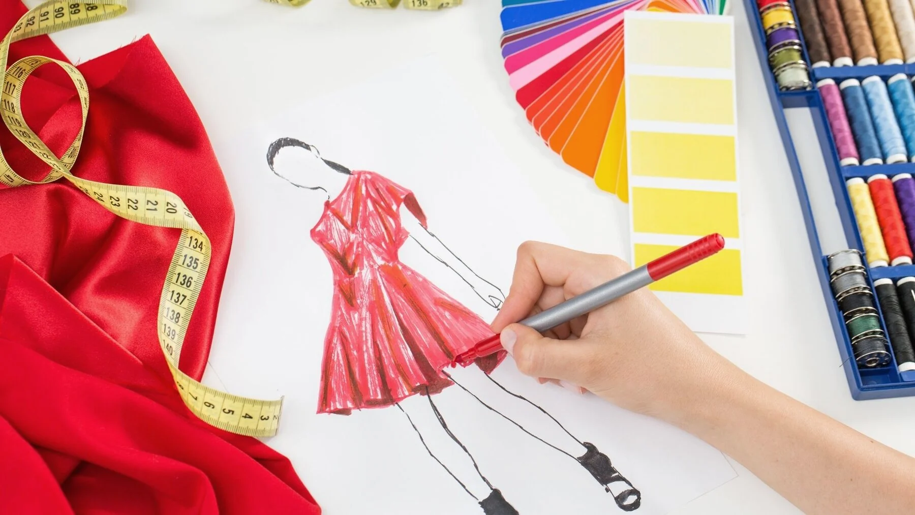 A fashion sketch of a red dress being coloured in, beside fabric, a tape measure and thread.