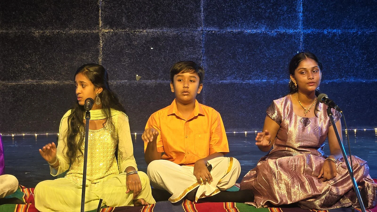 Three students singing Carnatic music on stage.